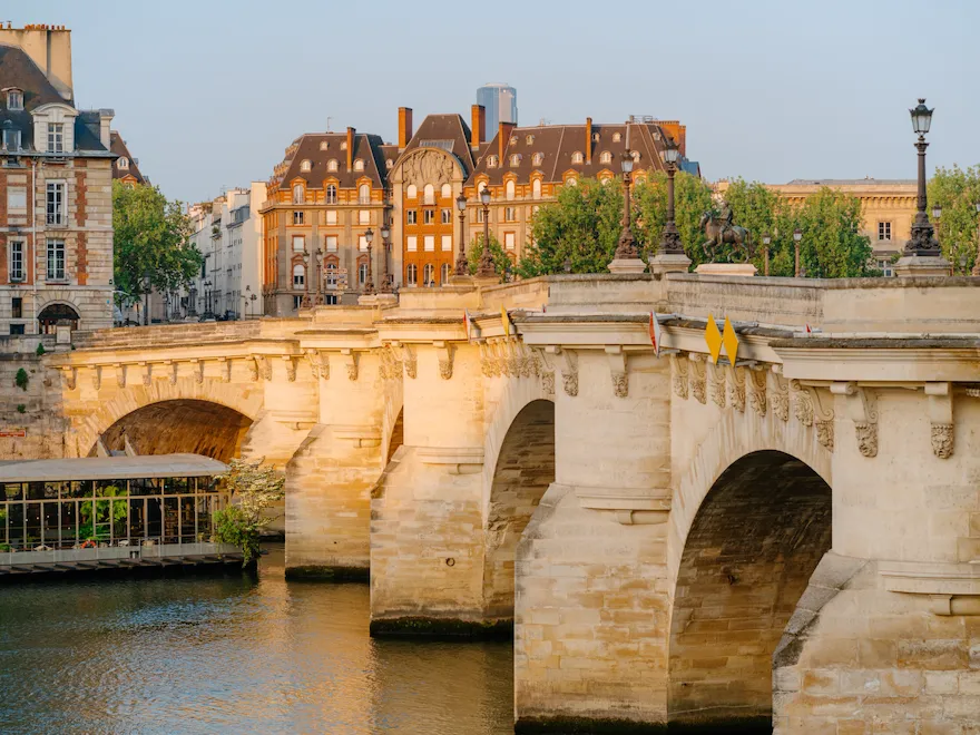 View of the Pont Neuf at sunrise, the Seine, and a boat filled with trees on the water, at Pont Neuf, Paris.