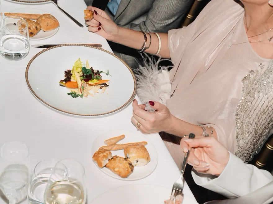 Detail of an event dinner with people eating and drinking red wine, with flowers detail in the foreground
