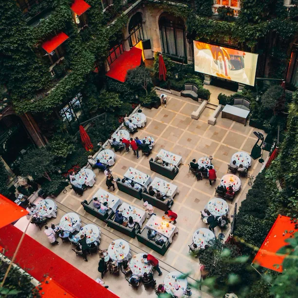 Vue aérienne d'un événement organisé dans une cour, où les invités sont assis à des tables rondes recouvertes de nappes blanches. Les touches de rouge et la verdure créent une atmosphère élégante.