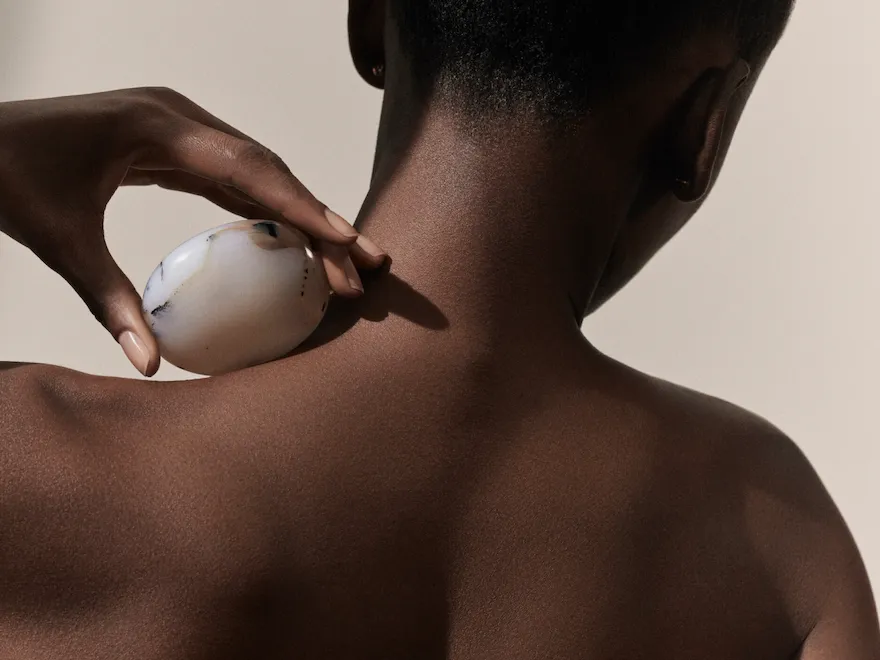 Dior Spa Plaza Athénée's iconography for spa treatments. Here, a dark-skin female model seen from the back and posing with a stone hold against the left-side of her neck.
