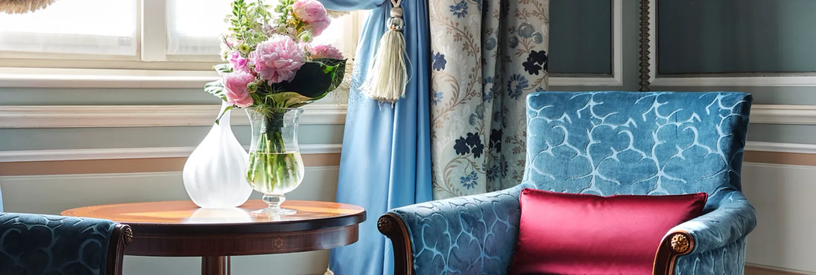 Dettaglio di una camera Mosaic, poltrona e tende sui toni dell'azzurro e fiori rosa sul tavolo sotto la finestra