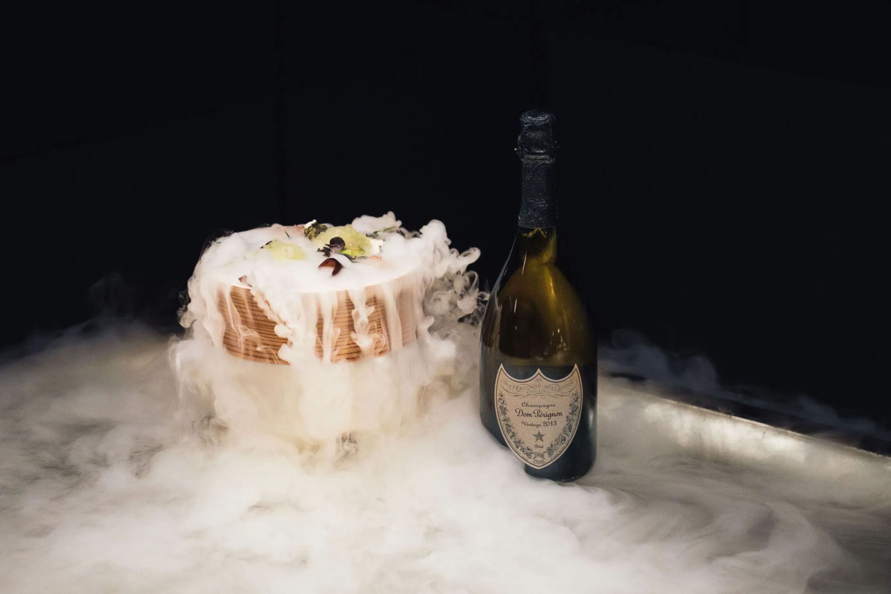 A decorated dessert emits smoke beside a bottle of Dom Pérignon champagne on a surface covered in mist.