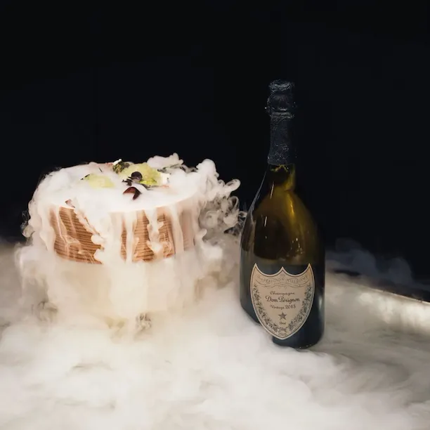 A decorated dessert emits smoke beside a bottle of Dom Pérignon champagne on a surface covered in mist.