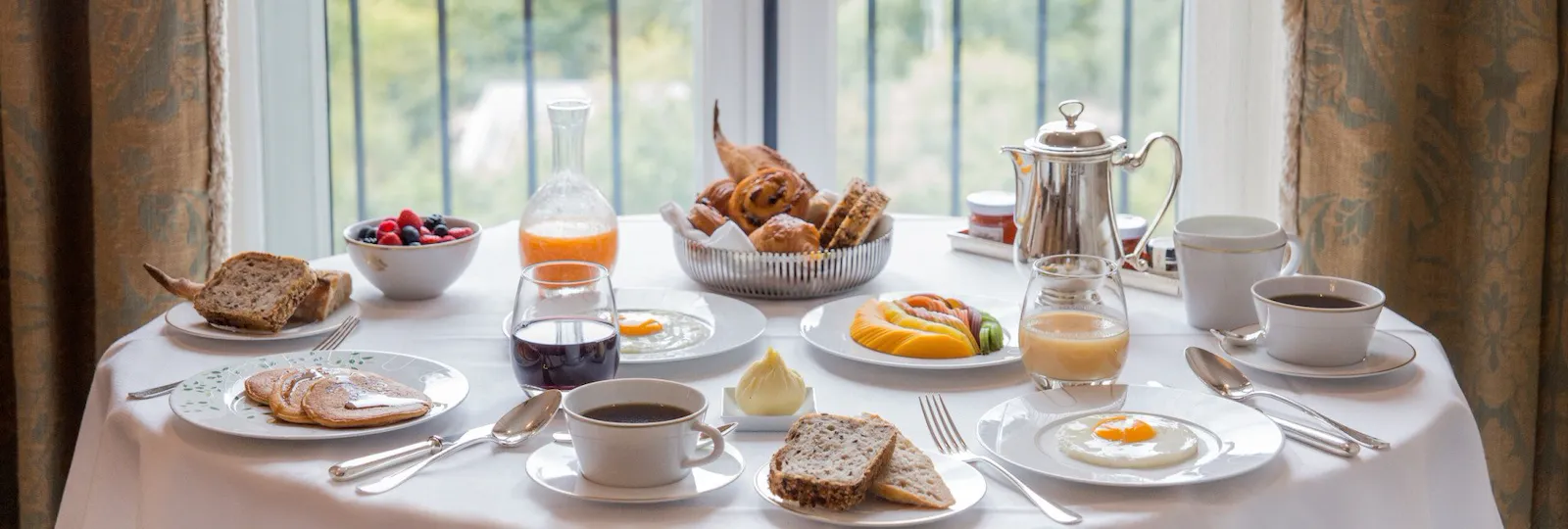 Table de room service dans son ensemble avec des aliments tels que les crêpes, les fruits, le pain et les pâtisseries et la vue lointaine du jardin des Tuileries à l’arrière. Au Meurice, à Paris.