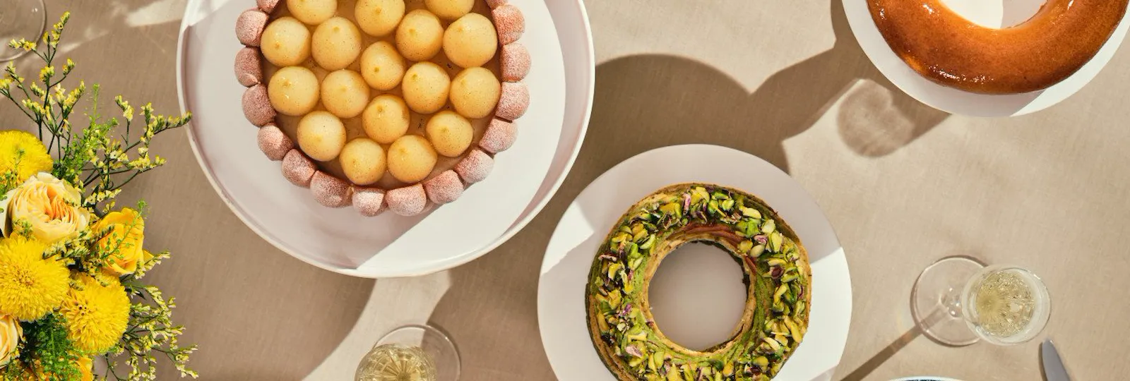 Uma mesa redonda com pratos elegantes, sobremesas e frutas. Flores amarelas estão dispostas num bouquet. O ambiente é acolhedor e convidativo, evocando um clima festivo.