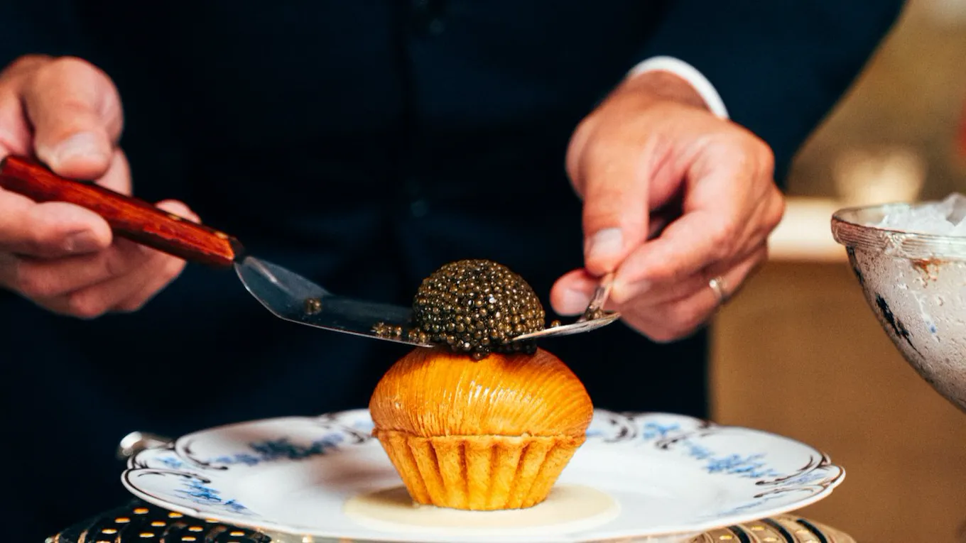A person slices a dessert topped with black caviar on a dinner plate in a formal setting.