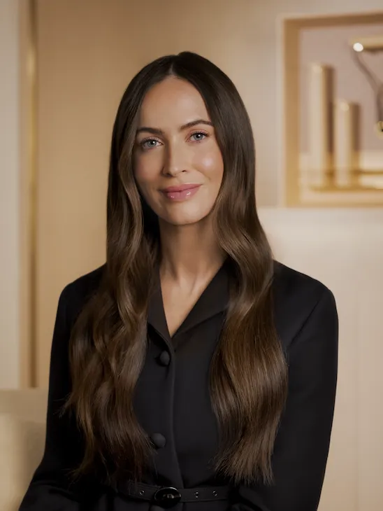 A woman smiling and sitting on a chair in a comfortable and warm room. She wears a black top.