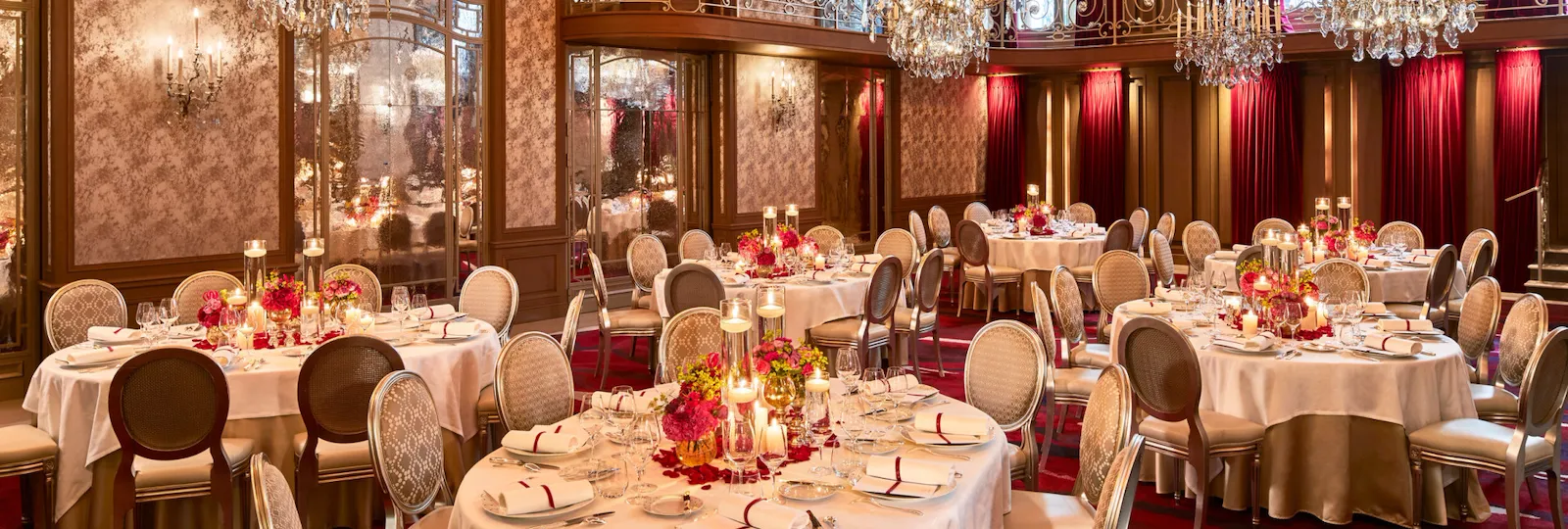 Elegante salão de banquetes com mesas redondas cobertas por toalhas brancas e decoradas com louças finas. Lustres de cristal e detalhes em vermelho criam um ambiente acolhedor.