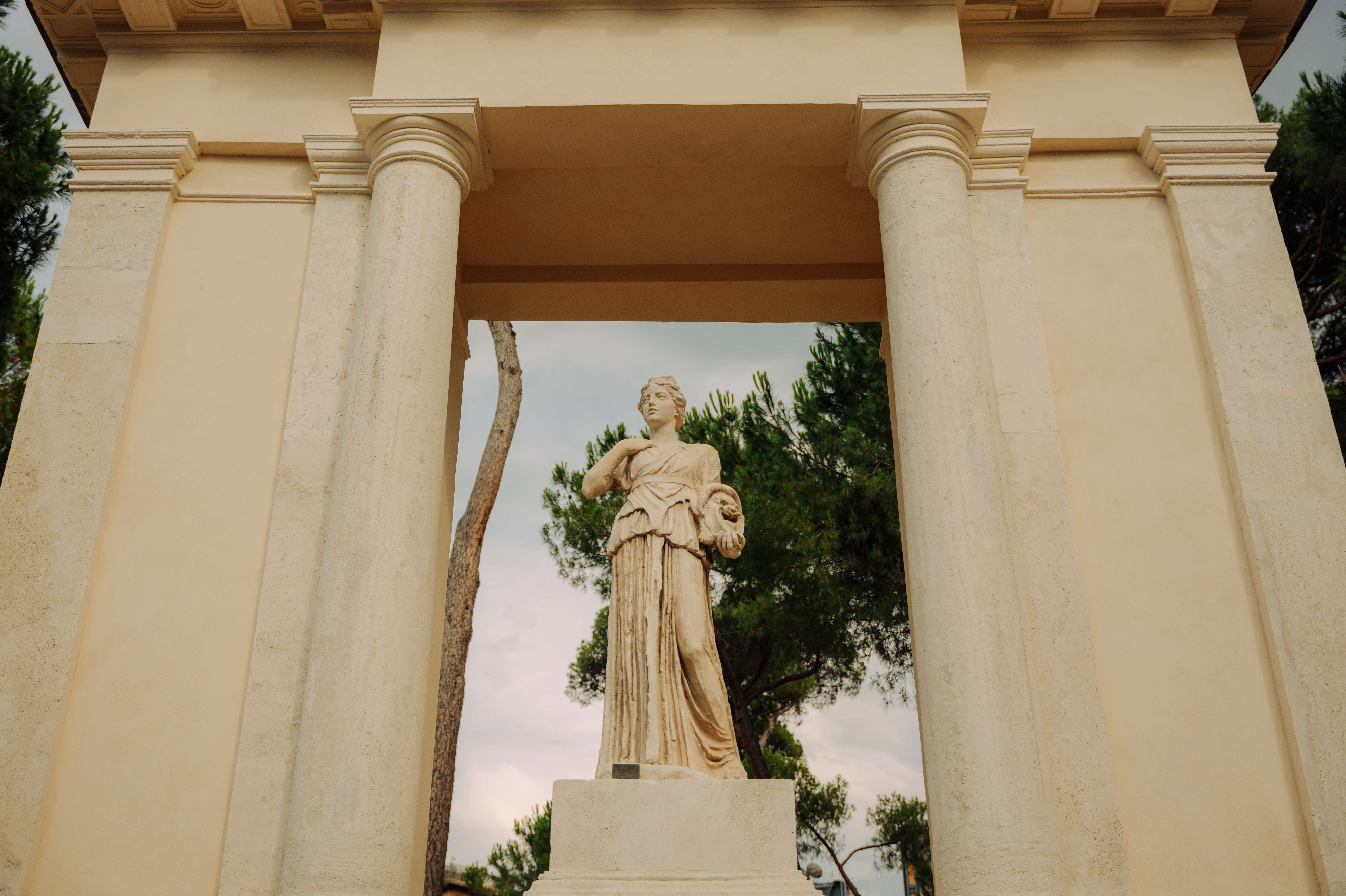 A statue of a figure standing in a classical setting with pillars and greenery behind