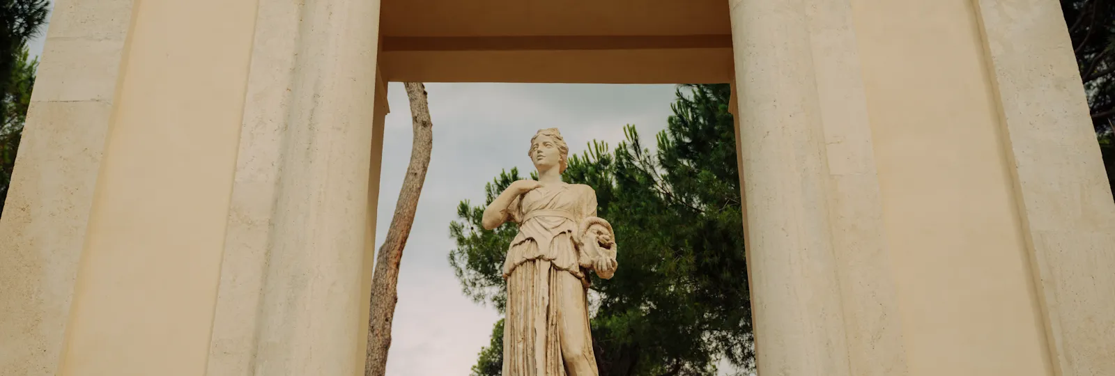 A statue of a figure standing in a classical setting with pillars and greenery behind