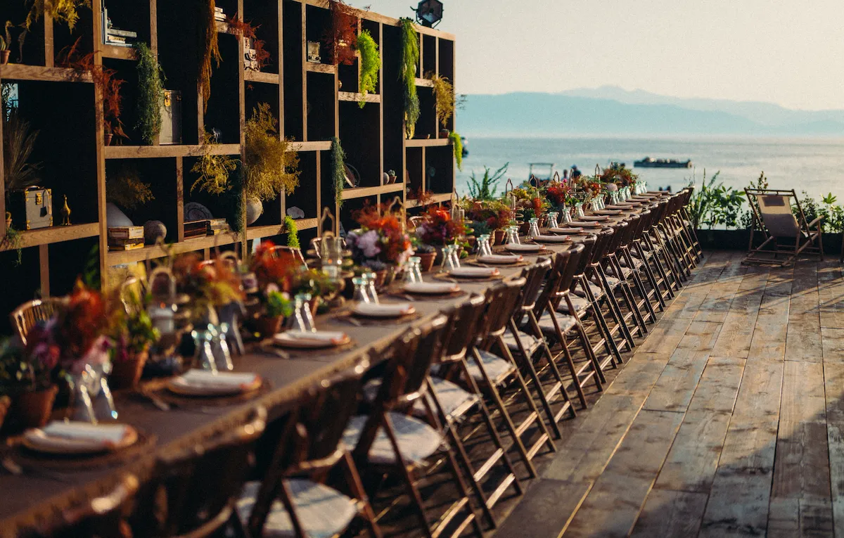 A round outdoor bar features tall wicker chairs and lush green and orange foliage. It stands on a wooden deck amid an inviting, sunlit garden setting.