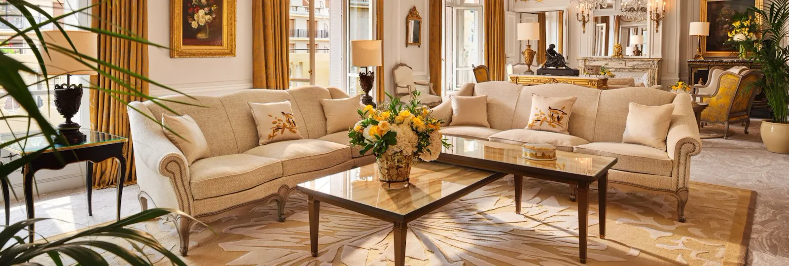Salon luxueux avec d'élégants canapés couleur crème, une table basse en verre et des touches dorées. Un lustre et de grandes fenêtres créent une ambiance lumineuse et opulente.
