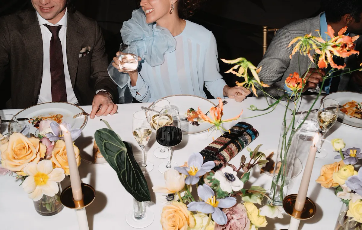 Detail of an event dinner with people eating and drinking red wine, with flowers detail in the foreground