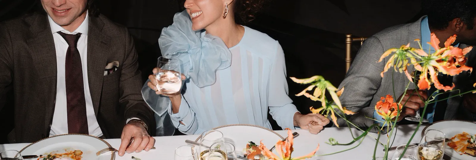 Detail of an event dinner with people eating and drinking red wine, with flowers detail in the foreground