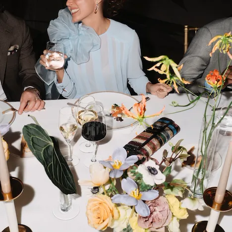 Detail of an event dinner with people eating and drinking red wine, with flowers detail in the foreground