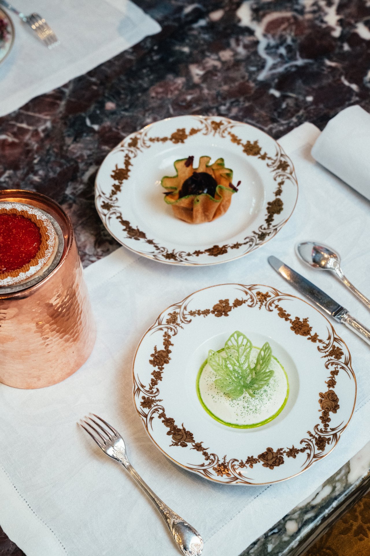 Elegant dining setup with decorative plates, small botanical-shaped entrees, silverware, and a copper tumbler.