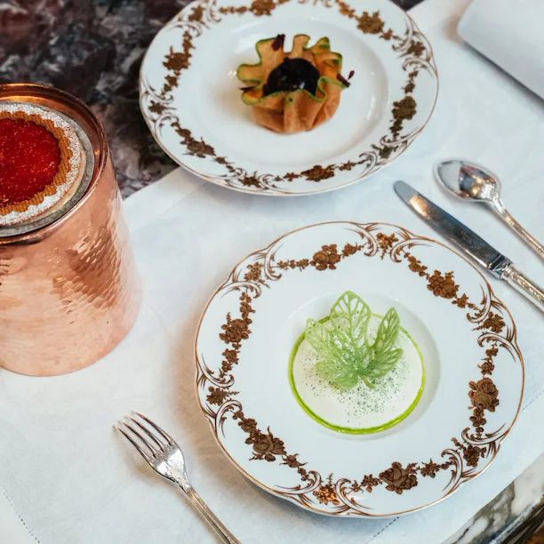 Elegant dining setup with decorative plates, small botanical-shaped entrees, silverware, and a copper tumbler.