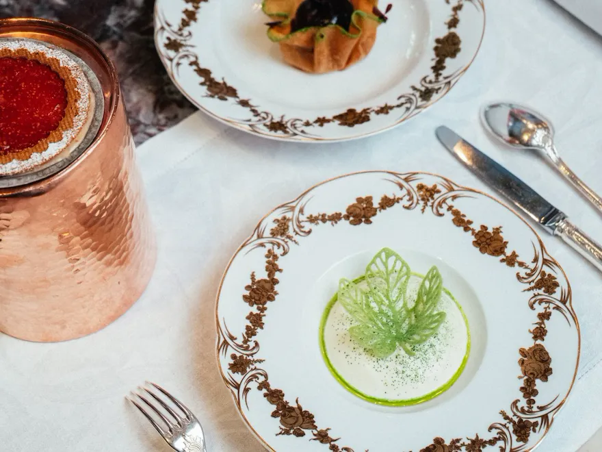 Elegant dining setup with decorative plates, small botanical-shaped entrees, silverware, and a copper tumbler.