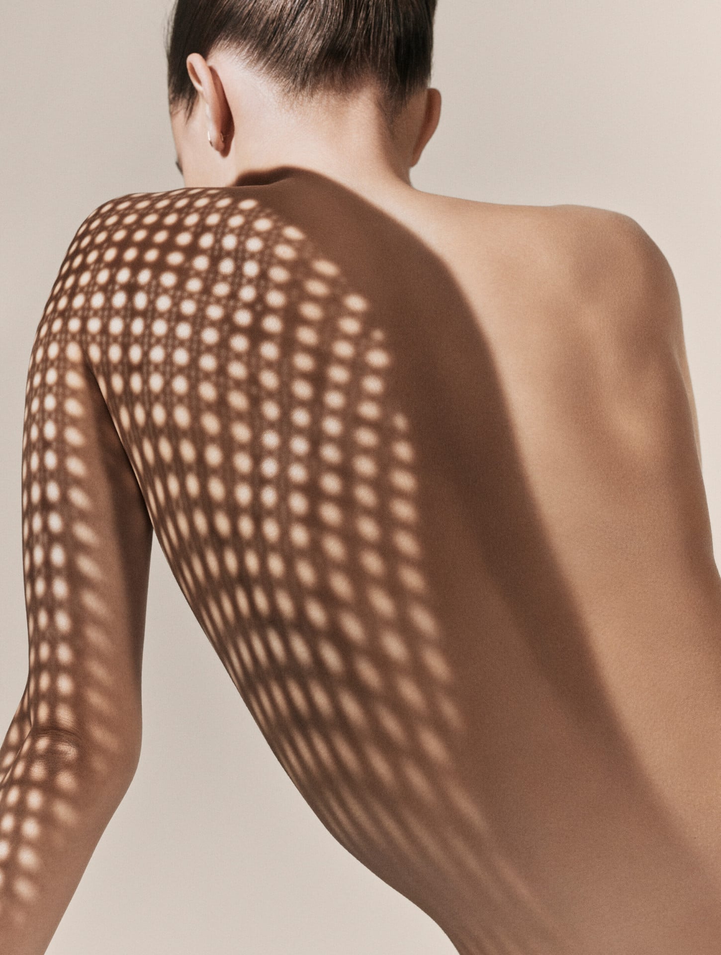 Dior Spa Plaza Athénée's iconography for spa treatments. Here, a light-skin female model with a hair bun posing naked with the shadow of a caning pattern chair on her back.