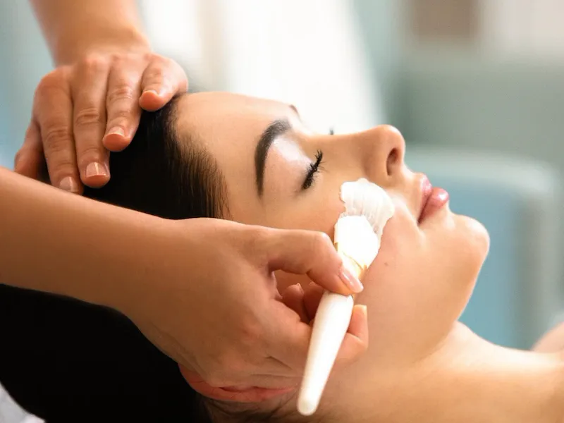 On this picture we can see a customer lying down, her head resting on a white pillow. A white treatment is applied to her face with a brush. The masseuse's other hand is placed on the client's hair. The client closes her eyes at Le Meurice, Paris.