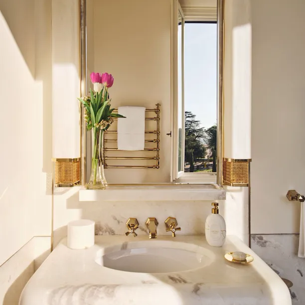 Detail of sink area with mirror reflecting the view from an open window in a guest room bathroom at Hotel Eden Rome
