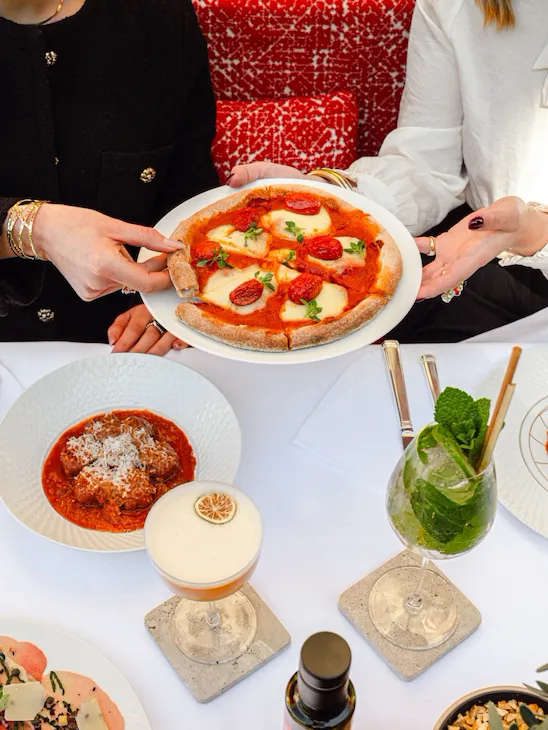 Due donne ad un tavolo con vari piatti e cocktail che condividono una pizza margherita