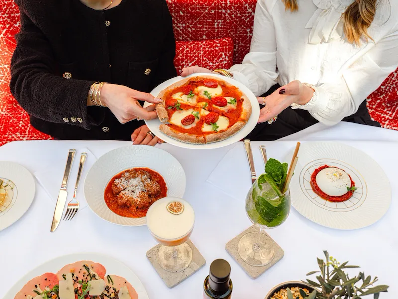 Due donne ad un tavolo con vari piatti e cocktail che condividono una pizza margherita