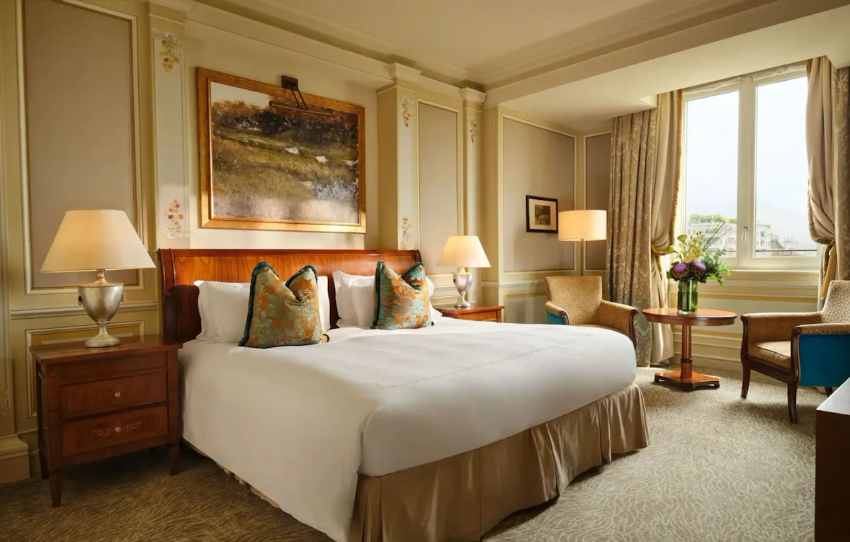 The room is decorated in soft gold colours and features decorative gold and blue cushions. There is a large painting above the bed and a large window that lets in plenty of natural light.