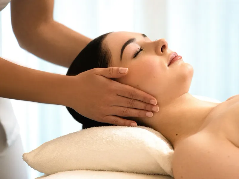 We can see a woman lying on a massage table, resting her head on a white pillow. We can see her from the side. The masseuse's hands are placed on the client's ears. The client's eyes are closed at the spa Valmont at Le Meurice, Paris.