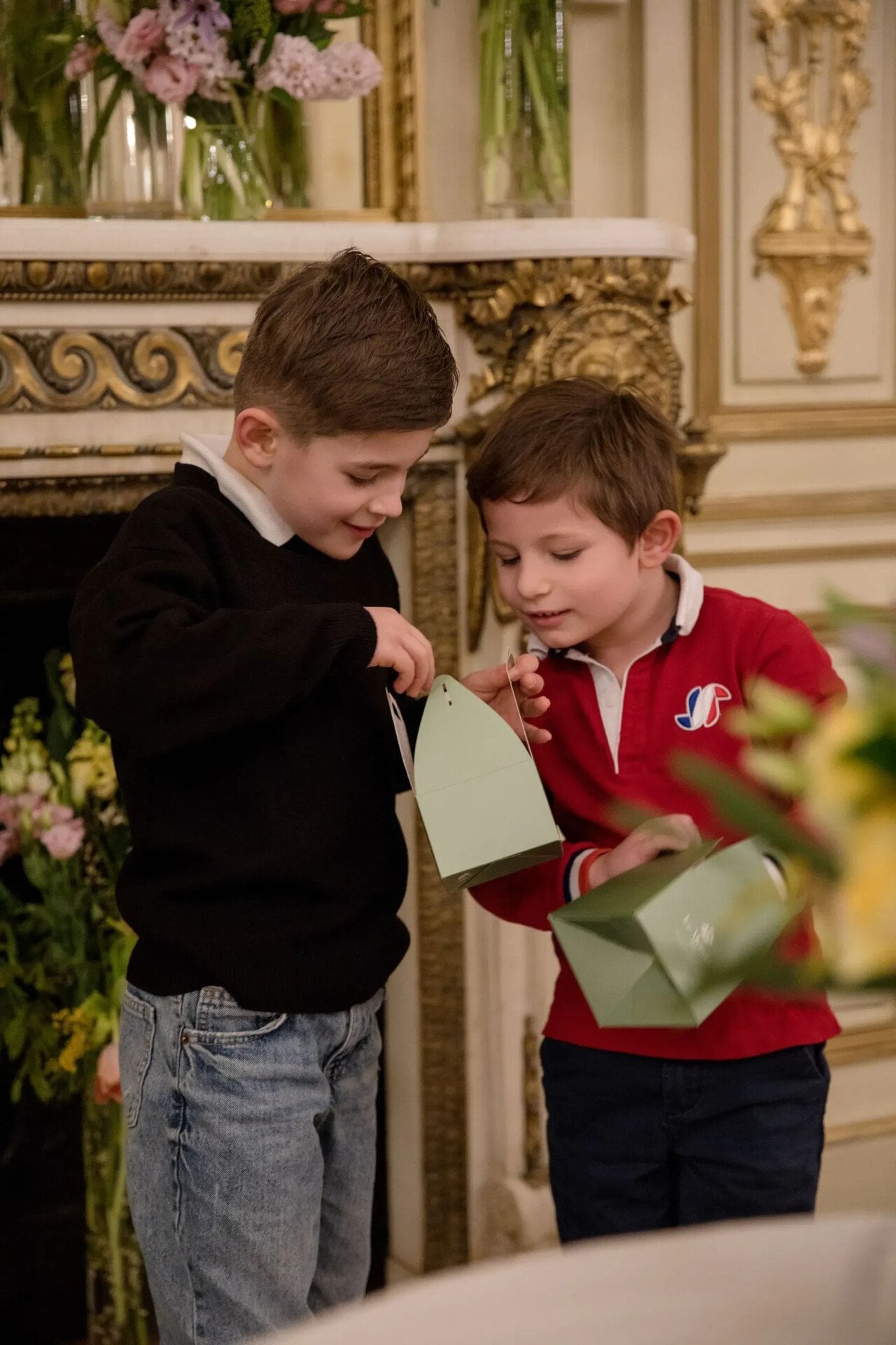 Two young boys stand in front of a fireplace, holding green boxes with handles, looking inside curiously. One wears a black sweater, the other a red polo. They are near an ornate fireplace with floral arrangements. At Le Meurice, Paris.