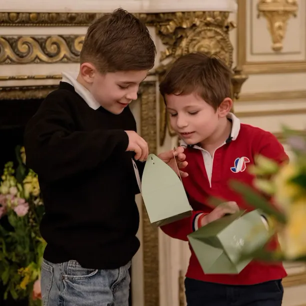 Two young boys stand in front of a fireplace, holding green boxes with handles, looking inside curiously. One wears a black sweater, the other a red polo. They are near an ornate fireplace with floral arrangements. At Le Meurice, Paris.