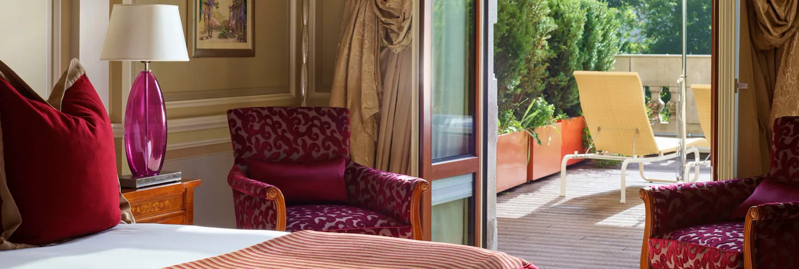 Vista della terrazza dalla camera da letto: portafinestra aperta, terrazza al sole con due sdraio e un ombrellone, in una giornata di sole