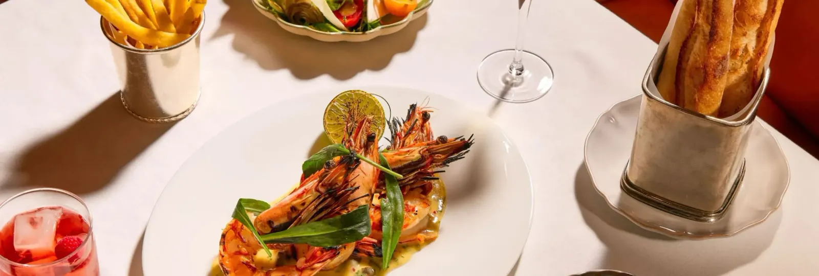 Dining table at The Grill at The Dorchester, showcasing grilled prawns served with herb butter and charred lime, accompanied by golden fries, a seasonal salad, green beans, fresh bread and a Martini cocktail.