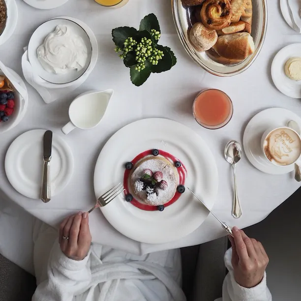 Vista dall' alto di un tavolo da servizio in camera con donna che mangia pancakes, yoghurt, succo, cappuccino e selezione di cornetti all' Hotel Eden.
