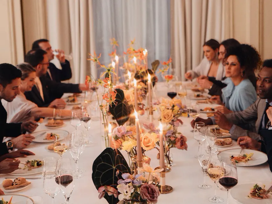 Cena con persone sedute intorno ad un tavolo imbandito e un agrande finestra sullo sfondo
