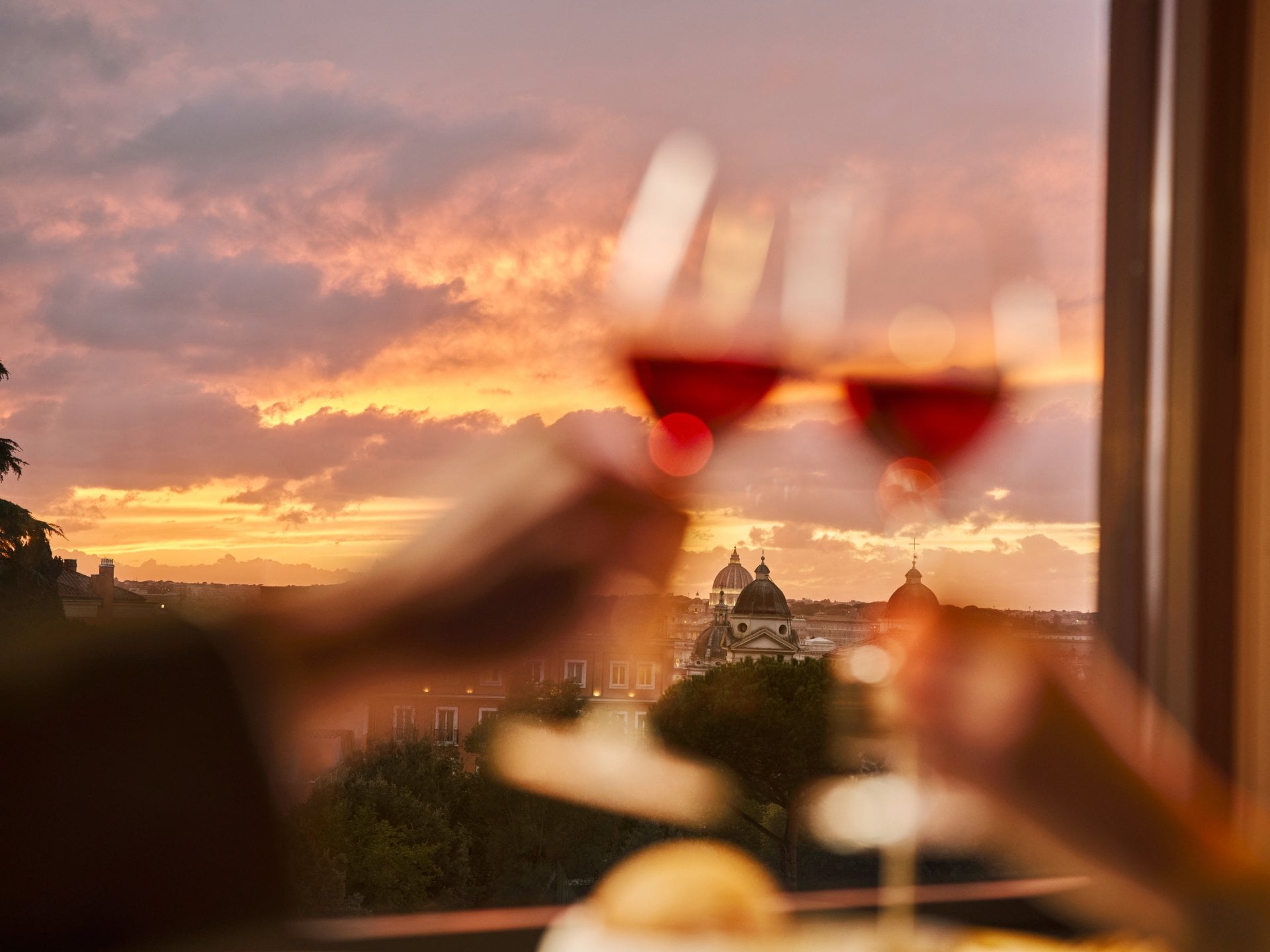 Two glasses of wine clink with a sunset view over a city skyline, creating a romantic mood.
