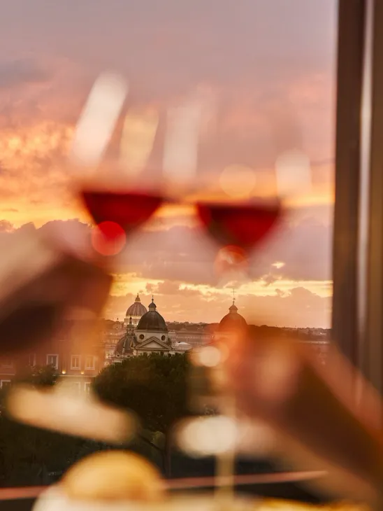 Due bicchieri di vino rossi avvicinati per un brindisi con lo sfondo di un tramonto su Roma