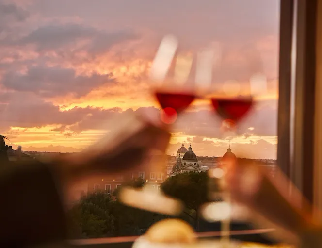 Two glasses of wine clink with a sunset view over a city skyline, creating a romantic mood.