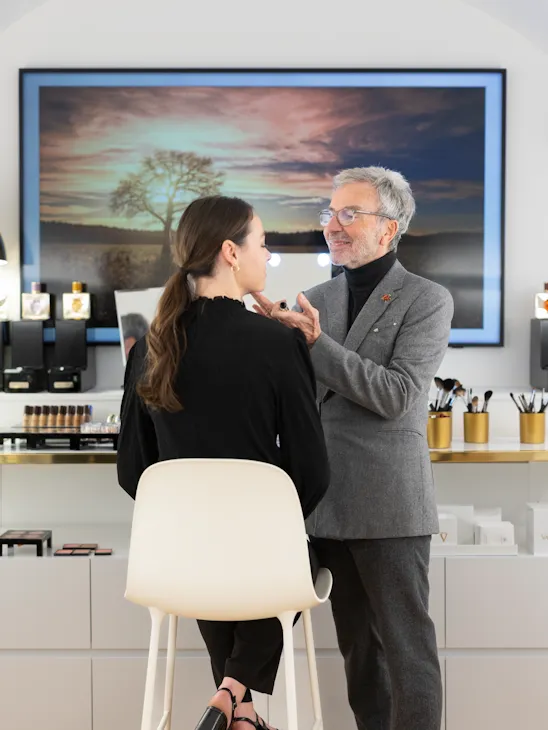A man in a gray suit is applying makeup to a woman seated in a white chair. They are in a modern makeup studio with beauty products and a large landscape photo in the background. The atmosphere is professional and focused.