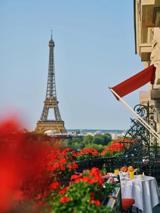 Vue depuis un balcon parisien avec la tour Eiffel au loin, entouré d'une végétation luxuriante. L'auvent rouge et les fleurs aux couleurs vives ajoutent chaleur et charme.