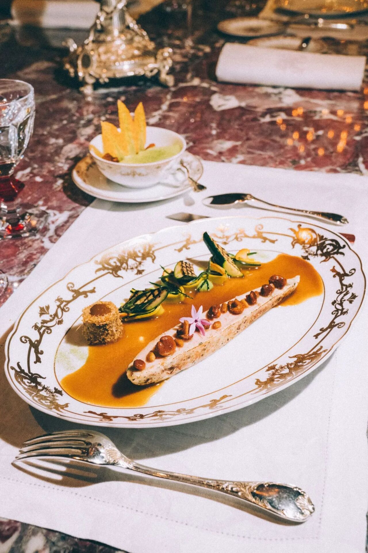 Elegant dish on ornate plate features chicken with sauce, small vegetables, and a flower garnish. Cup with crackers nearby on a luxurious table setting.