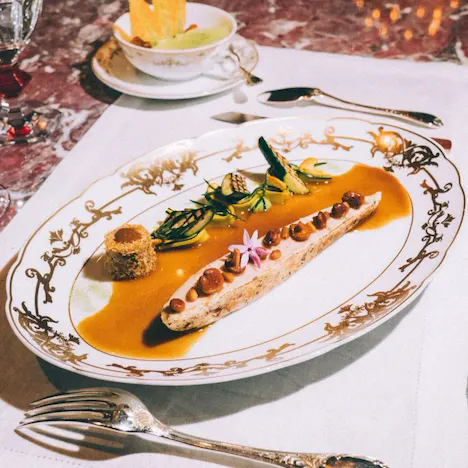 Elegant dish on ornate plate features chicken with sauce, small vegetables, and a flower garnish. Cup with crackers nearby on a luxurious table setting.