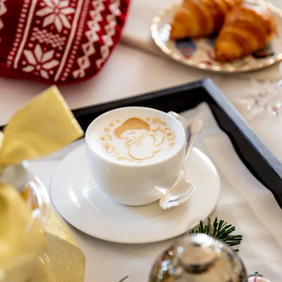 Cappuccino con immagine di Babbo Natale, cornetti, calza della befana e decorazioni natalizie disposte su di una tovaglia