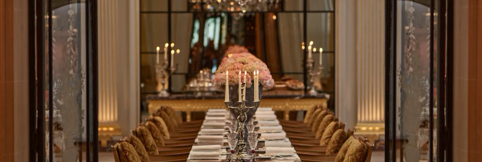 La table centrale en marbre du Jean Imbert au Plaza Athénée, décorée de fleurs roses et de lustres. Des lustres en cristal suspendus au-dessus de la table. Une porte en fer forgé et en verre au premier plan. Hôtel Plaza Athénée, Paris.