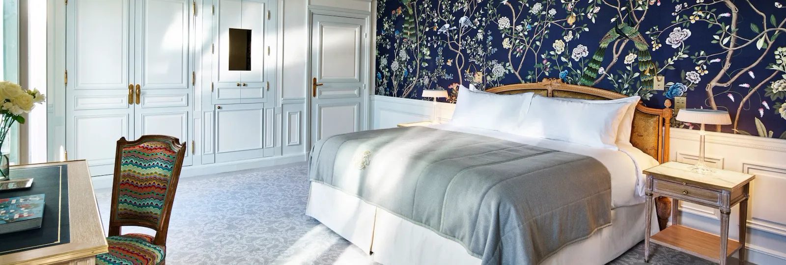 Une chambre luxueuse avec un grand lit recouvert d'une couette grise et d'oreillers blancs. Le papier peint bleu à motifs floraux ajoute une touche d'élégance. Les armoires blanches et la chaise à motifs renforcent l'impression de luminosité et d'espace de la pièce. Une lumière douce pénètre dans la pièce, créant une atmosphère sereine.