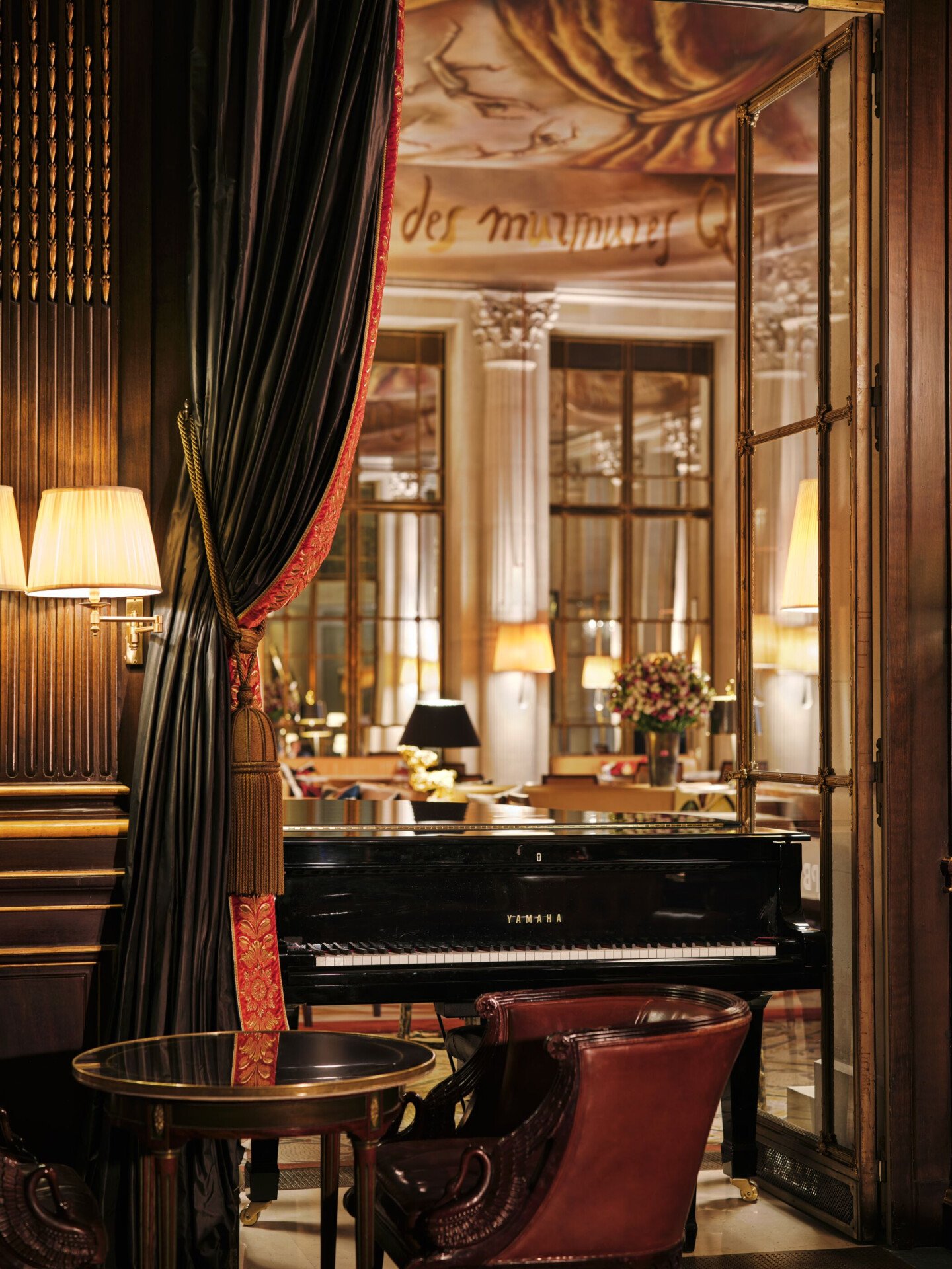 View from an English bar with wooden walls on a black piano. In the background you can see a restaurant with a brown toned canva on the ceiling. In the front of the picture you can see a round table with a leather chair. The piano is placed in a window door which is open. A heavy curtain with trimmings frames the window door, at Le Meurice, Paris