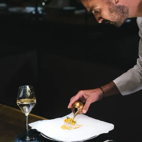 A chef is plating a delicate dish with precision using a brass tool. A glass of white wine sits on the table, creating an elegant and refined atmosphere.