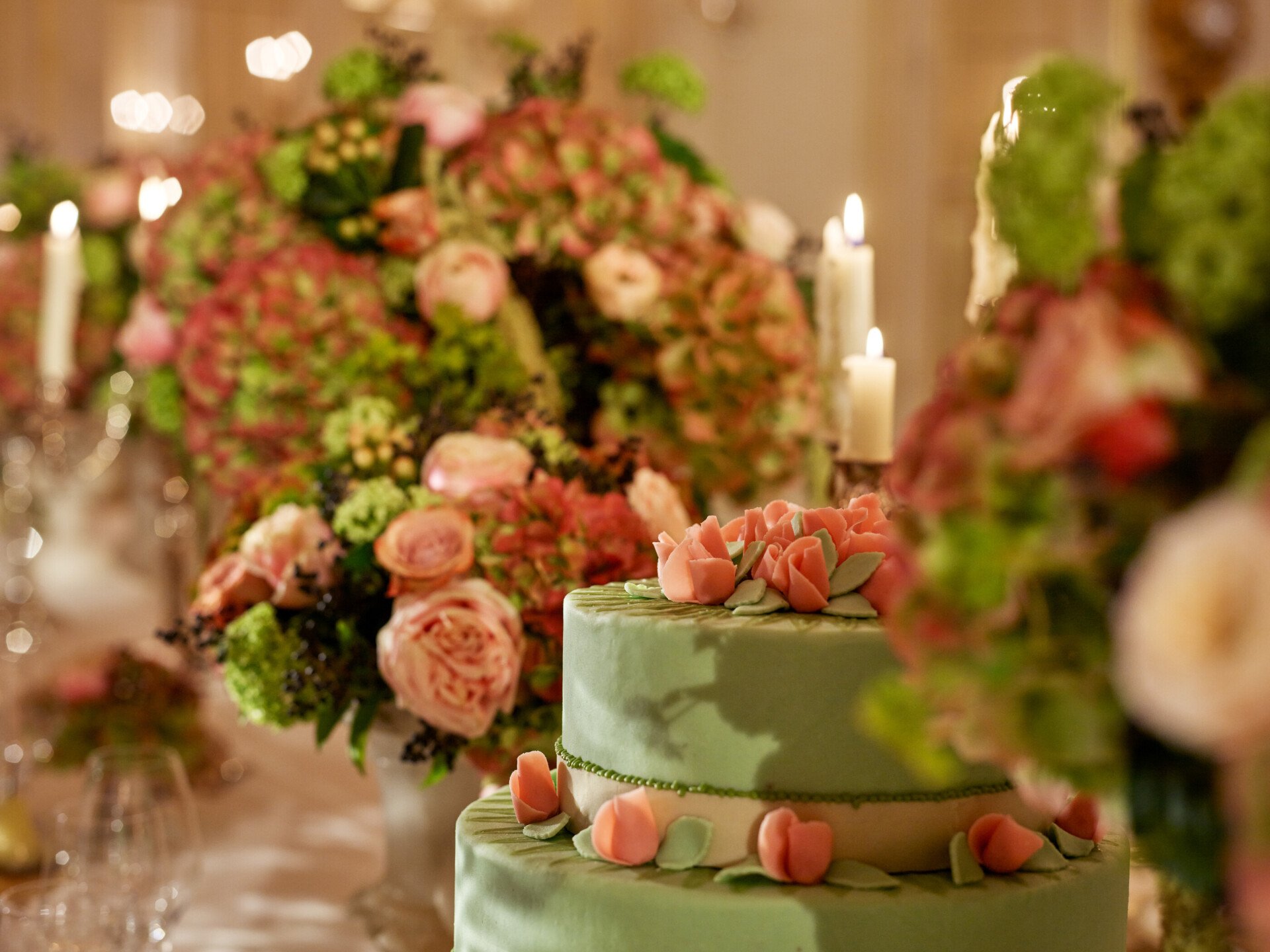 Élégant gâteau vert orné de roses roses, entouré de somptueux arrangements floraux et de bougies allumées. La scène dégage une atmosphère romantique et festive.