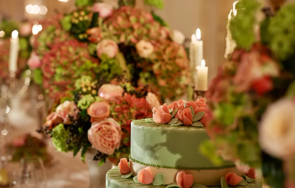 Élégant gâteau vert orné de roses roses, entouré de somptueux arrangements floraux et de bougies allumées. La scène dégage une atmosphère romantique et festive.