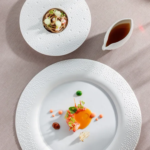 Two round tables laid ona table one with a langoustine on a sauce and the other with ramen in it, next a bowl with ramen broth